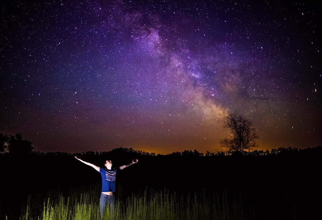 A woman standing quietly at a window at night, looking up at a star-filled sky