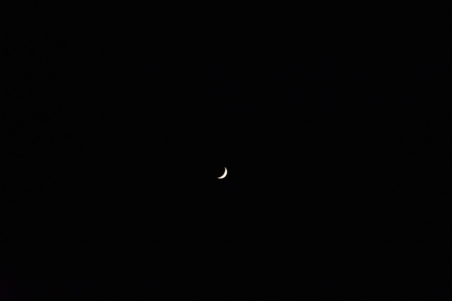 A thin new moon crescent glowing in a deep indigo night sky above quiet water